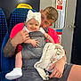 baby, man, baby_carrier, bow, tattoo, train_seat, train_interior, smile, white_shoes, knitted_blanket, pacifier, shorts, pink_shirt, window, door, sign, person, child, indoor, transport