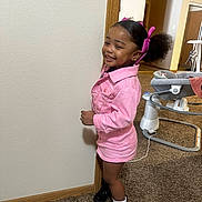 Lucy is registered to the contest to win money with this photo: child, girl, pink_jacket, hair_bows, black_boots, white_socks, carpet, indoor, baby_walker, smiling, standing, wall, doorway, room, hair, happy, person, footwear, floor, clothing
