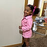 child, girl, pink_jacket, hair_bows, black_boots, white_socks, carpet, indoor, baby_walker, smiling, standing, wall, doorway, room, hair, happy, person, footwear, floor, clothing
