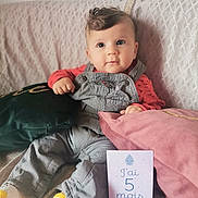 Esser a rejoint le concours — aidez-le/la à gagner de superbes lots ! baby, infant, child, couch, pillow, clothing, overalls, socks, milestone_card, text, face, cute, indoor, portrait, seated, soft_furnishing, curly_hair, looking_away, cozy, happy