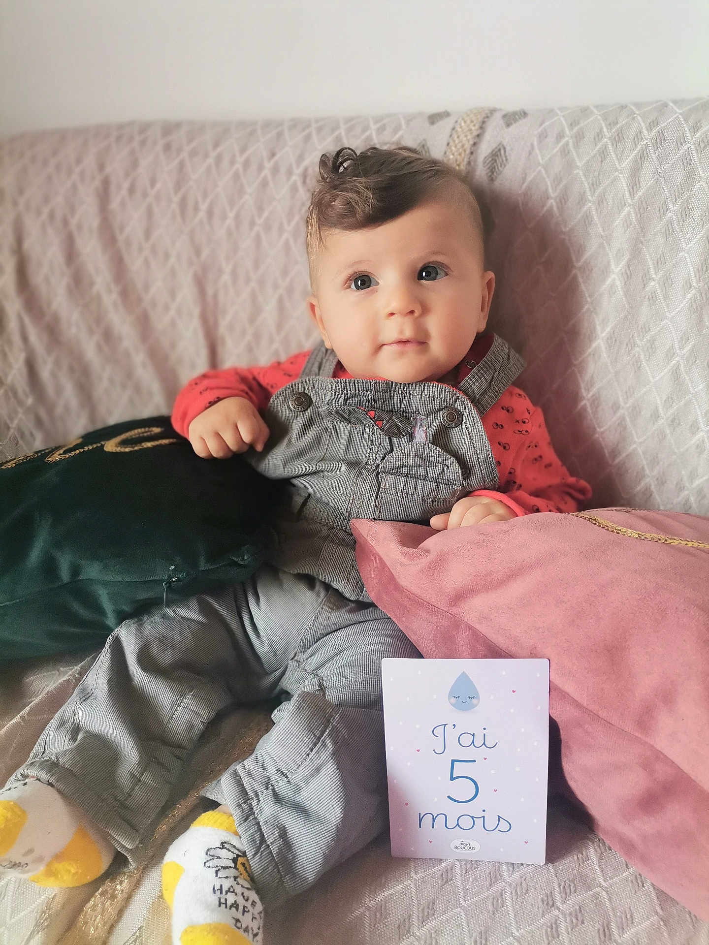 Esser a rejoint le concours — aidez-le/la à gagner de superbes lots ! baby, infant, child, couch, pillow, clothing, overalls, socks, milestone_card, text, face, cute, indoor, portrait, seated, soft_furnishing, curly_hair, looking_away, cozy, happy