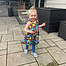 child, girl, smiling, dress, floral_pattern, leggings, sneakers, outdoor, patio, stone_floor, furniture, brick_wall, happy, walking, daylight, curly_hair, playful, person, backyard, sunny