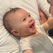 Theodore is registered to the contest to win money with this photo: baby, infant, smiling, laughing, onesie, bed, pillow, portrait, close_up, happy_face, joyful, parent, interaction, lying_down, expression, eyes, mouth, hand, skin, bedding