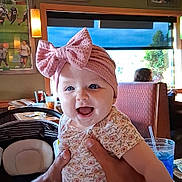 Scarlett joined the competition — help win amazing prizes! baby, child, pink_bow, headband, floral_outfit, restaurant, booth, table, glass, straw, window, greenery, poster, sports, hands, smile, happy, indoor, person, infant