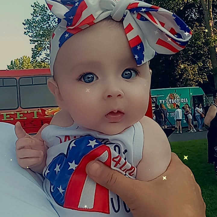 Scarlett is registered to the contest to win money with this photo: baby, bow, child, clothing, cute, daytime, face, festival, food_truck, hand, headband, holding, outdoor, park, patriotic, people, person, sparkles, summer, trees