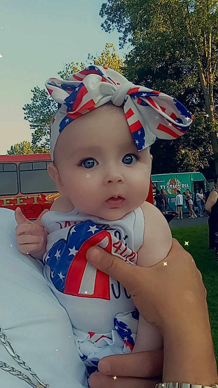 Scarlett is registered to the contest to win money with this photo: baby, child, face, headband, bow, outdoor, person, patriotic, clothing, hand, food_truck, trees, summer, festival, cute, holding, people, park, daytime, sparkles
