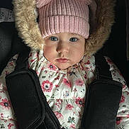 Alma joined the competition — help win amazing prizes! baby, child, car_seat, winter_jacket, pink_hat, pom_poms, floral_pattern, blue_eyes, face, hands, clothing, straps, safety_harness, cute, indoors, portrait, infant, warm_clothing, fur_trim, seat