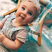 Izya a rejoint le concours — aidez-le/la à gagner de superbes lots ! baby, blonde_hair, blue_eyes, candid, casual_clothing, child, close_up, curious, face, floor, happy, natural_light, outdoor, overalls, person, portrait, short_sleeves, smiling, toddler, wooden_chair