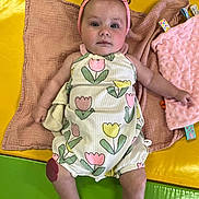 Isabella is registered to the contest to win money with this photo: baby, infant, child, headband, romper, floral_pattern, blanket, toy, yellow_mat, green_mat, pink_blanket, lying_down, indoors, cute, soft_texture, colorful, portrait, skin, face, expression