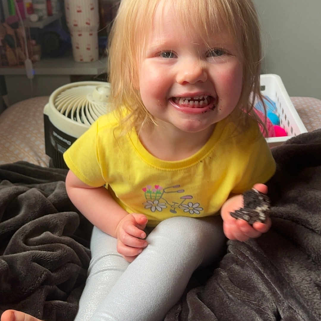 Sawyer is registered to the contest to win money with this photo: toddler, child, smiling, cookie, yellow_shirt, leggings, blanket, barefoot, indoor, fan, furniture, person, happy, face, hair, room, toy, background, sitting, cozy