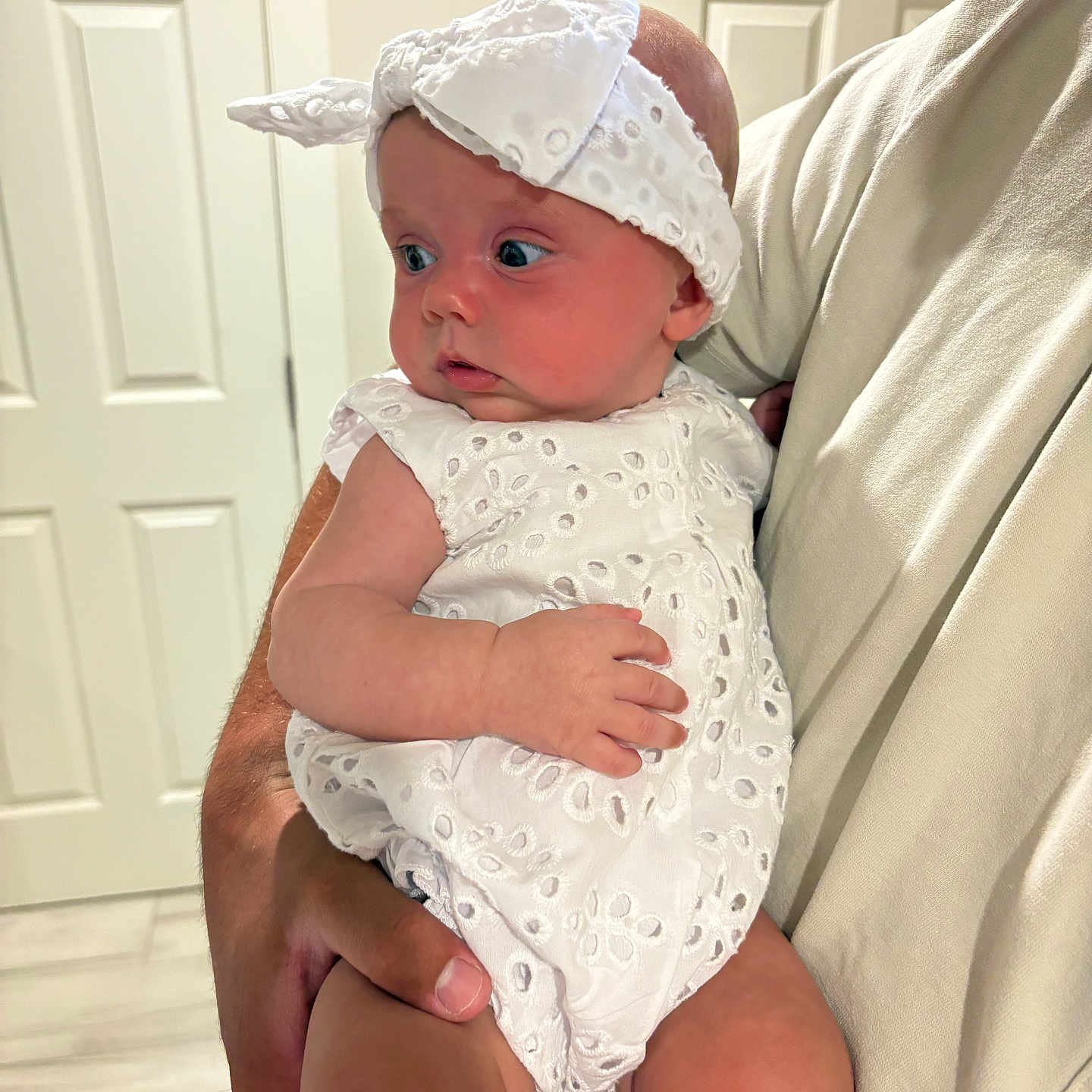 Scottie is registered to the contest to win money with this photo: adult_hand, baby, child, close_up, curious, cute, door, expression, floor, headband, holding, indoor, infant, looking_away, person, portrait, skin, white_dress, white_shirt, young_child