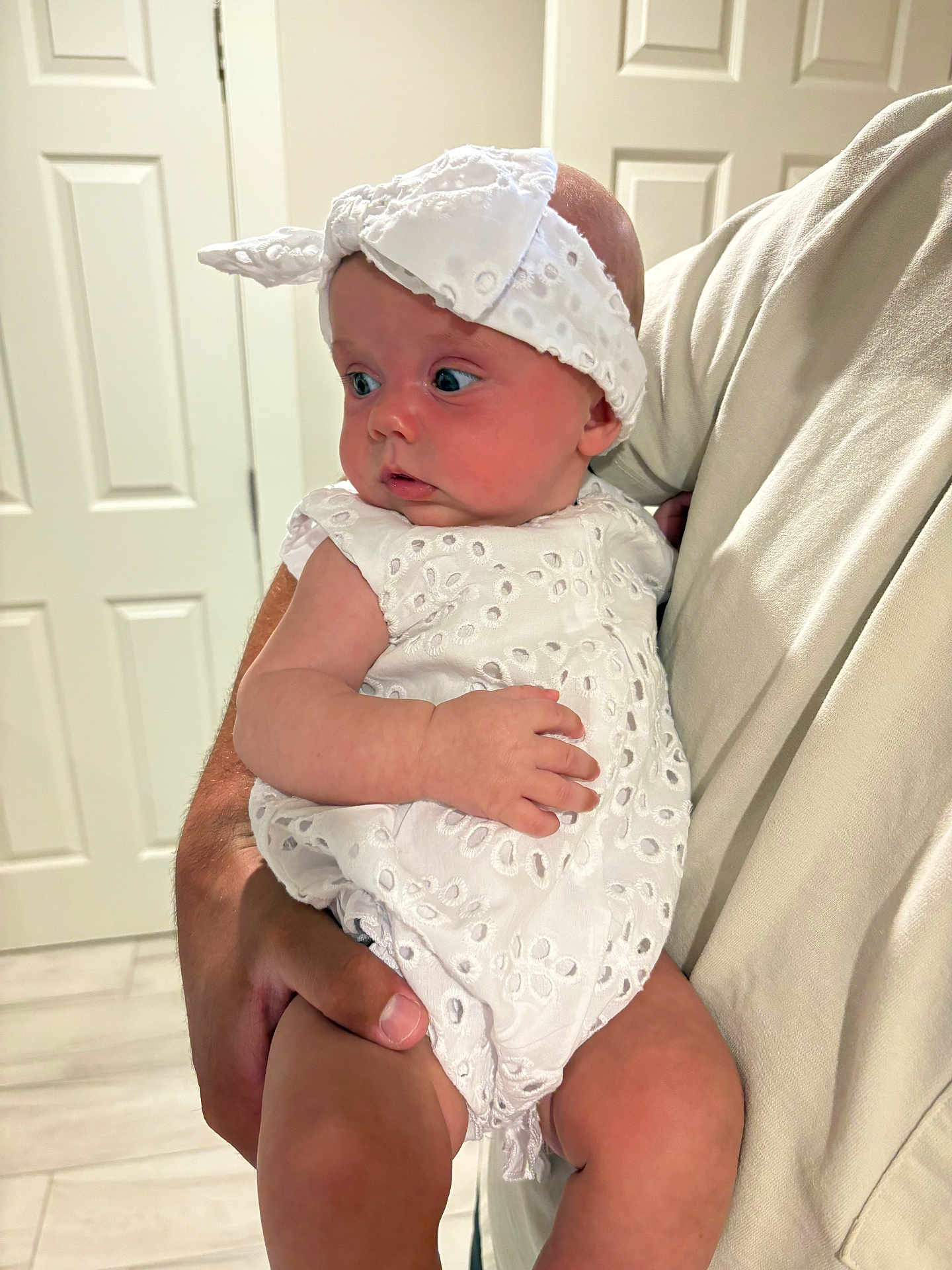 Scottie is registered to the contest to win money with this photo: baby, infant, child, white_dress, headband, adult_hand, indoor, curious, looking_away, white_shirt, holding, skin, door, floor, portrait, close_up, person, young_child, cute, expression