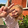 child, smiling, peace_sign, outdoor, brick_wall, green_plants, sunlight, happy, casual_clothing, playful, portrait, daylight, person, young_girl, nature, concrete_ground, long_hair, gesture, cheerful, cute
