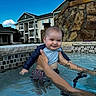 baby, infant, child, smiling, swimming_pool, water, outdoor, summer, adult, hands, tattoo, stone_wall, fountain, apartment_building, blue_sky, clouds, portrait, family, daylight, happy