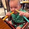 baby, child, toddler, smiling, eating, restaurant, high_chair, spoon, toy, green_outfit, short_hair, portrait, close_up, indoor, dining, table, chair, happy, candid, safe