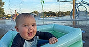 Ezekiel is registered to the contest to win money with this photo: baby, child, infant, smiling, happy, outdoor, splash_pad, water, inflatable_pool, swimwear, wet_hair, water_drops, fountain, summer, sunny, portrait, playground, recreation, pool, face