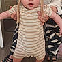 baby, child, person, hat, striped_clothing, onesie, indoor, floor_mat, animal_pattern, barefoot, standing, supporting_hands, adult_hands, footwear, wooden_floor, home_interior, expression, curious, cute, playful