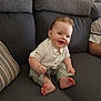 baby, smiling, sitting, couch, gray_couch, pillow, striped_pillow, white_shirt, khaki_pants, barefoot, feet, chubby_cheeks, happy, indoor, portrait, candid, timestamp, watermark, person, home