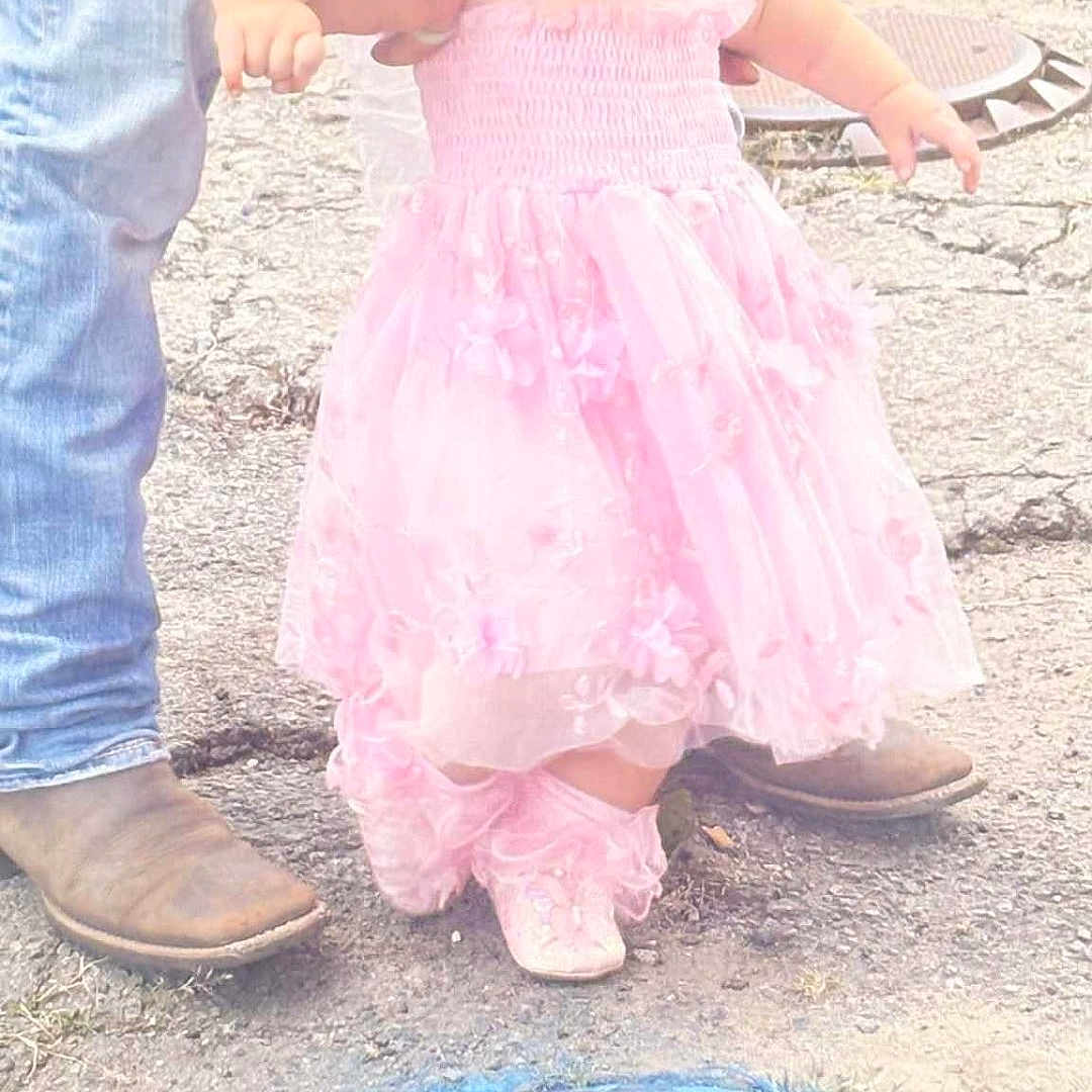 Scarlett is registered to the contest to win money with this photo: adult_hand, baby_shoes, boots, casual_clothing, child, curious, floral_headband, flower_accessory, footwear, helping_hand, jeans, manhole_cover, outdoor, pavement, pink_dress, sidewalk, street, support, toddler, walking