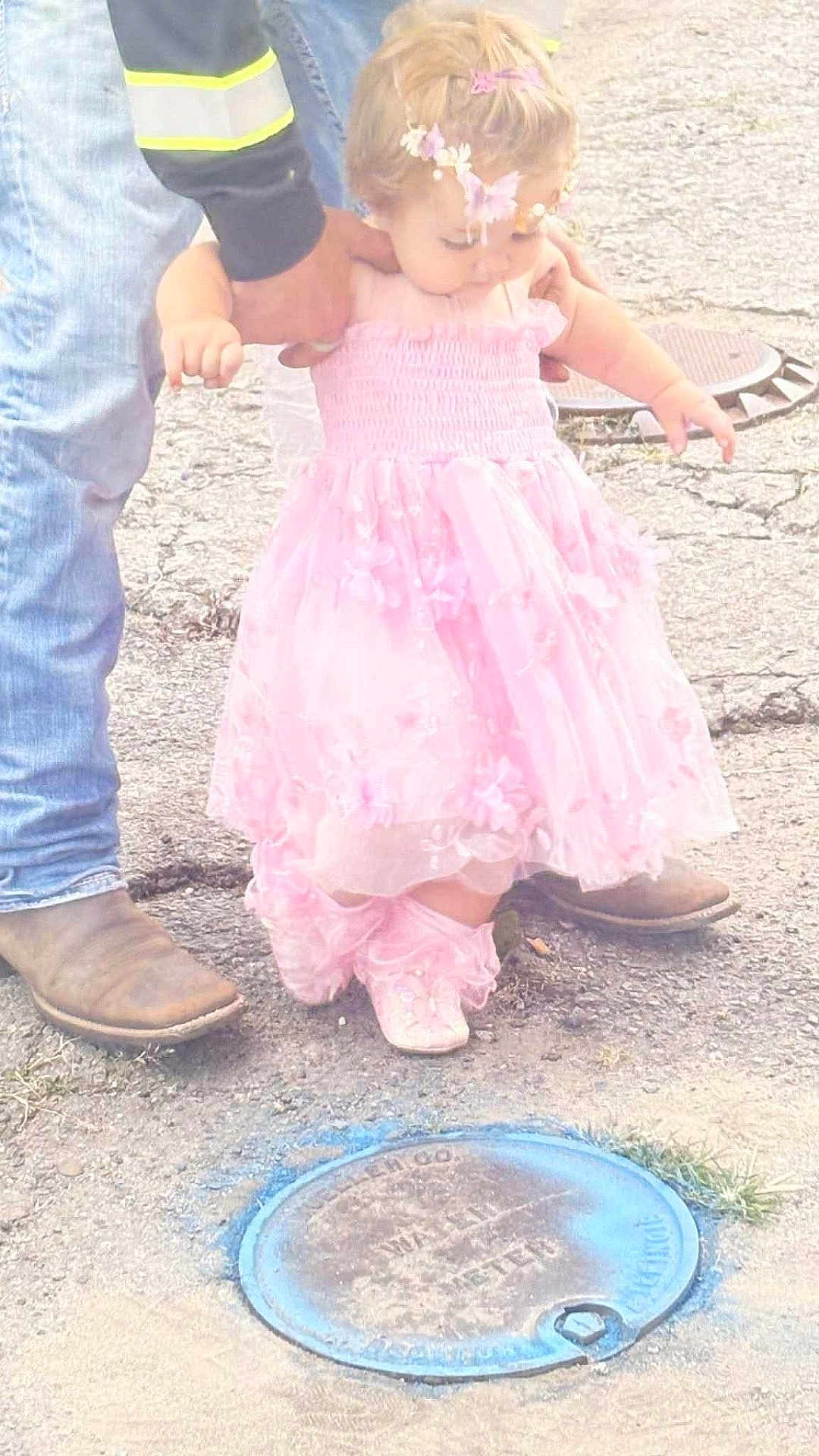 Scarlett is registered to the contest to win money with this photo: child, toddler, pink_dress, floral_headband, boots, jeans, adult_hand, pavement, manhole_cover, outdoor, walking, support, curious, footwear, flower_accessory, casual_clothing, baby_shoes, street, helping_hand, sidewalk