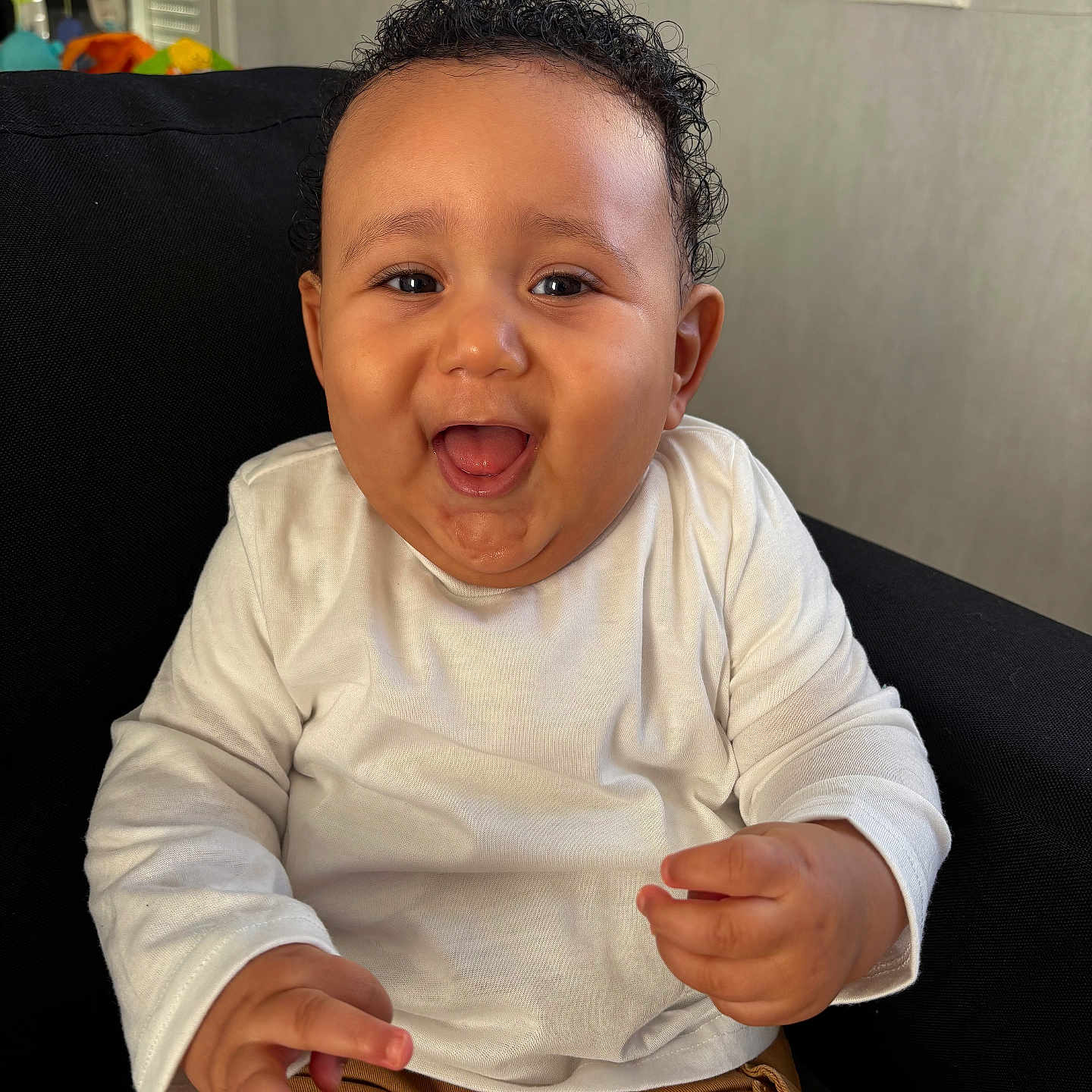 Izaak a rejoint le concours — aidez-le/la à gagner de superbes lots ! baby, black_chair, brown_pants, child, clothing, curly_hair, cute, expression, face, hands, happy, indoor, person, portrait, seated, smiling, toddler, white_shirt, window, young_child
