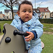 Izaak a rejoint le concours — aidez-le/la à gagner de superbes lots ! blue_onesie, child, curly_hair, daytime, expression, face, grass, hand, houses, outdoor, park, person, playground, rocker, sky, toddler, toy, tree, urban, winter_clothing