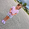child, girl, standing, street, sidewalk, cloudy_sky, suburban, residential_area, shorts, tshirt, purple_clothing, shoes, crocs, holding_object, daylight, asphalt, trees, houses, curious_expression, outdoor