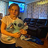 casual_clothing, child, curtains, dark_pants, family_room, home, hugging, indoor, living_room, playful, plush_toy, portrait, recliner, sitting, smiling, sofa, stitch, t_shirt, television, wooden_floor