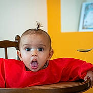 Olivia participe au concours pour gagner de l'argent avec cette photo : baby, child, high_chair, red_bib, spoon, food, surprised, expression, indoor, wooden_chair, person, hair_tie, hand, feeding, mouth_open, wide_eyes, face, cute, infant, colorful_background