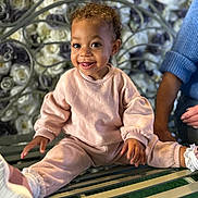 Malika joined the competition — help win amazing prizes! toddler, child, smiling, pink_clothing, bench, flower_wall, curly_hair, happy, casual_wear, indoor, person, adult_arm, green_bench, white_shoes, decorative_background, portrait, cute, seated, playful, cozy