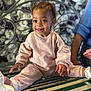 toddler, child, smiling, pink_clothing, bench, flower_wall, curly_hair, happy, casual_wear, indoor, person, adult_arm, green_bench, white_shoes, decorative_background, portrait, cute, seated, playful, cozy