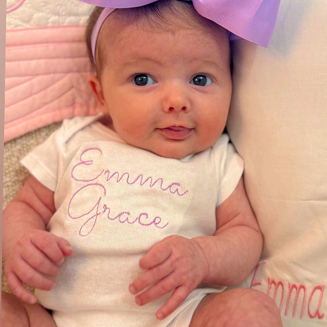 Emma Grace is registered to the contest to win money with this photo: baby, bow, child, clothing, cute, face, hands, headband, infant, legs, newborn, onesie, pillow, pink, portrait, purple, quilt, smiling, soft_texture, text_embroidery