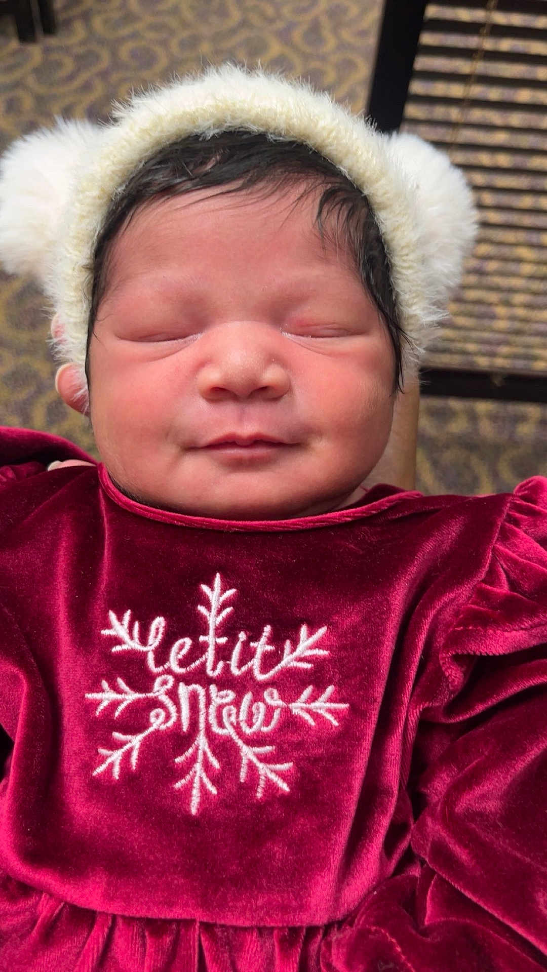 Elizah is registered to the contest to win money with this photo: baby, newborn, infant, sleeping, face, hat, pom_pom, velvet_dress, burgundy, embroidery, snowflake, closeup, portrait, cozy, chubby_cheeks, dark_hair, seated, indoor, patterned_carpet, subtle_smile