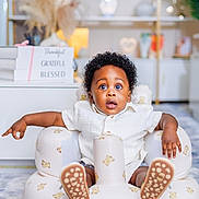 Zayan joined the competition — help win amazing prizes! baby, blessed, blurred_background, carpet, child, curly_hair, cute, grateful, home_decor, indoor, infant, inflatable_chair, shelf, sign, sitting, surprised_expression, teddy_bear_pattern, thankful, white_shirt, wide_eyes