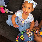 Paris is registered to the contest to win money with this photo: toddler, child, bow_headband, blue_dress, tutu, couch, marvel_comics, sippy_cup, indoor, person, happy, climbing, toy, necklace, hand, fabric, patterned_clothing, playful, cute, young_child