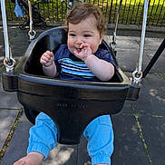 Aiden is registered to the contest to win money with this photo: baby, child, swing, playground, outdoor, park, smiling, thumb_sucking, blue_pants, black_swing, sunlight, shadow, fence, stroller, person, casual_clothing, curly_hair, barefoot, happy, daytime