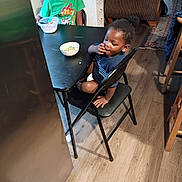 Da'Twan is registered to the contest to win money with this photo: child, children, indoor, kitchen, table, chair, cereal, bowl, floor, wood_floor, person, furniture, casual, snack, young, curly_hair, green_shirt, blue_shirt, eating, home
