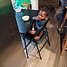 child, children, indoor, kitchen, table, chair, cereal, bowl, floor, wood_floor, person, furniture, casual, snack, young, curly_hair, green_shirt, blue_shirt, eating, home