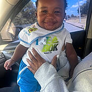 Mack is registered to the contest to win money with this photo: baby, child, smiling, portrait, car, car_interior, passenger, adult_hand, bib, t_shirt, blue_shorts, white_clothing, sitting, standing, outdoor_view, sky, trees, road, sunlight, happy