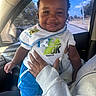 baby, child, smiling, portrait, car, car_interior, passenger, adult_hand, bib, t_shirt, blue_shorts, white_clothing, sitting, standing, outdoor_view, sky, trees, road, sunlight, happy