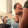 baby, infant, child, smiling, curly_hair, brown_skin, adult, parent, holding, family, indoor, home, floor, wooden_floor, dog, pet, slippers, jeans, portrait, candid