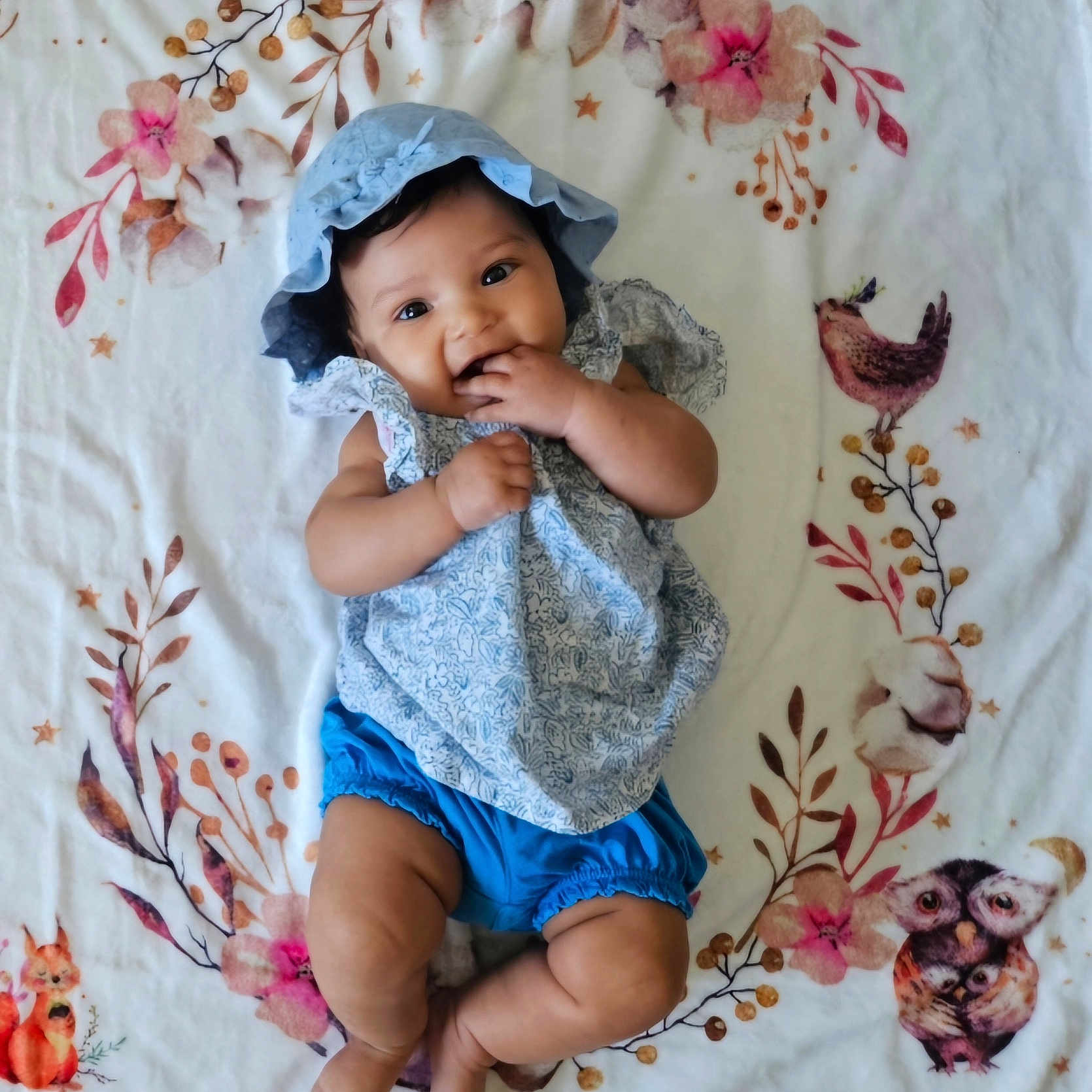 Mia a rejoint le concours — aidez-le/la à gagner de superbes lots ! adorable, animal_pattern, baby, blanket, blue_bonnet, blue_shorts, bright_eyes, chewing_fingers, child, cute, floral_pattern, hands, happy, indoors, infant, legs, lying_down, portrait, smiling, soft_texture