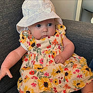 Alice is registered to the contest to win money with this photo: baby, bodypart, bonnet, cap, clothing, coat, couch, dress, eating, face, finger, food, furniture, hat, head, person, photography, portrait, sitting, sunhat