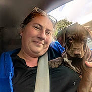 Scarlett is registered to the contest to win money with this photo: blue_jacket, brown_dog, car_interior, casual_clothing, close_up, daylight, dog, glasses, happy, holding, outdoor_view, pet, portrait, puppy, seatbelt, short_hair, smile, trees, window, woman