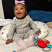 Jade is registered to the contest to win money with this photo: baby, infant, smiling, headband, pink_bow, floral_outfit, sitting, pillow, toy, rattle, socks, playtime, carpet, indoor, cute, hand, feet, cheerful, portrait, dark_background