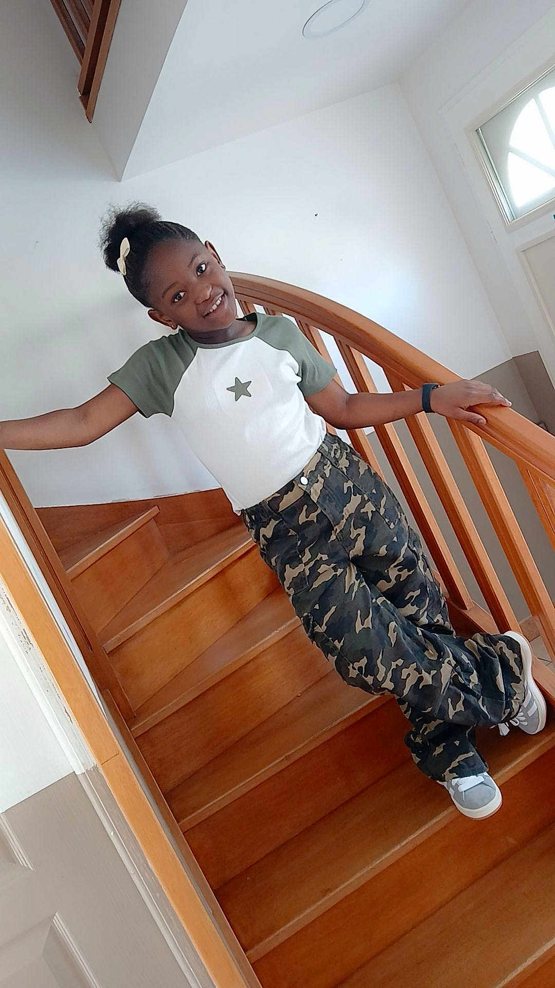 Kendra participe au concours pour gagner de l'argent avec cette photo : child, girl, smile, stairs, wooden_stairs, railing, indoor, casual_clothing, camouflage_pants, tshirt, hair_bow, sneakers, happy, portrait, person, home, handrail, watch, young, posing