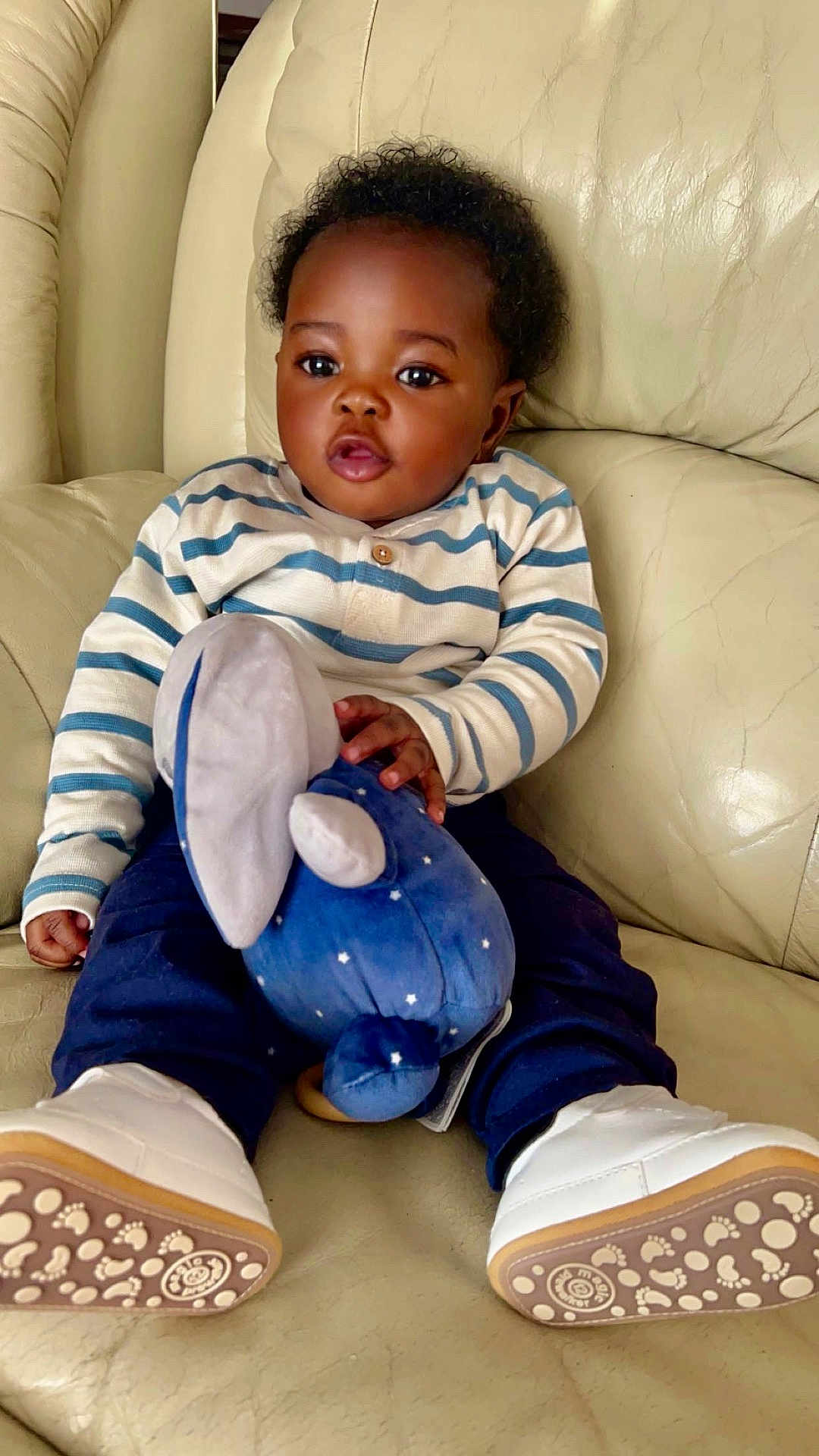 Dany a rejoint le concours — aidez-le/la à gagner de superbes lots ! baby, child, striped_shirt, navy_pants, white_shoes, plush_toy, stuffed_animal, couch, leather, indoor, sitting, cute, curious, smiling, footwear, toy, person, infant, cozy, home
