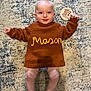 Mason joined the competition — help win amazing prizes! baby, infant, child, knitted_sweater, brown_sweater, name_on_clothing, milestone_marker, rug, floor, barefoot, smiling, lying_down, cute, happy, person, young_child, indoors, portrait, cozy, playful