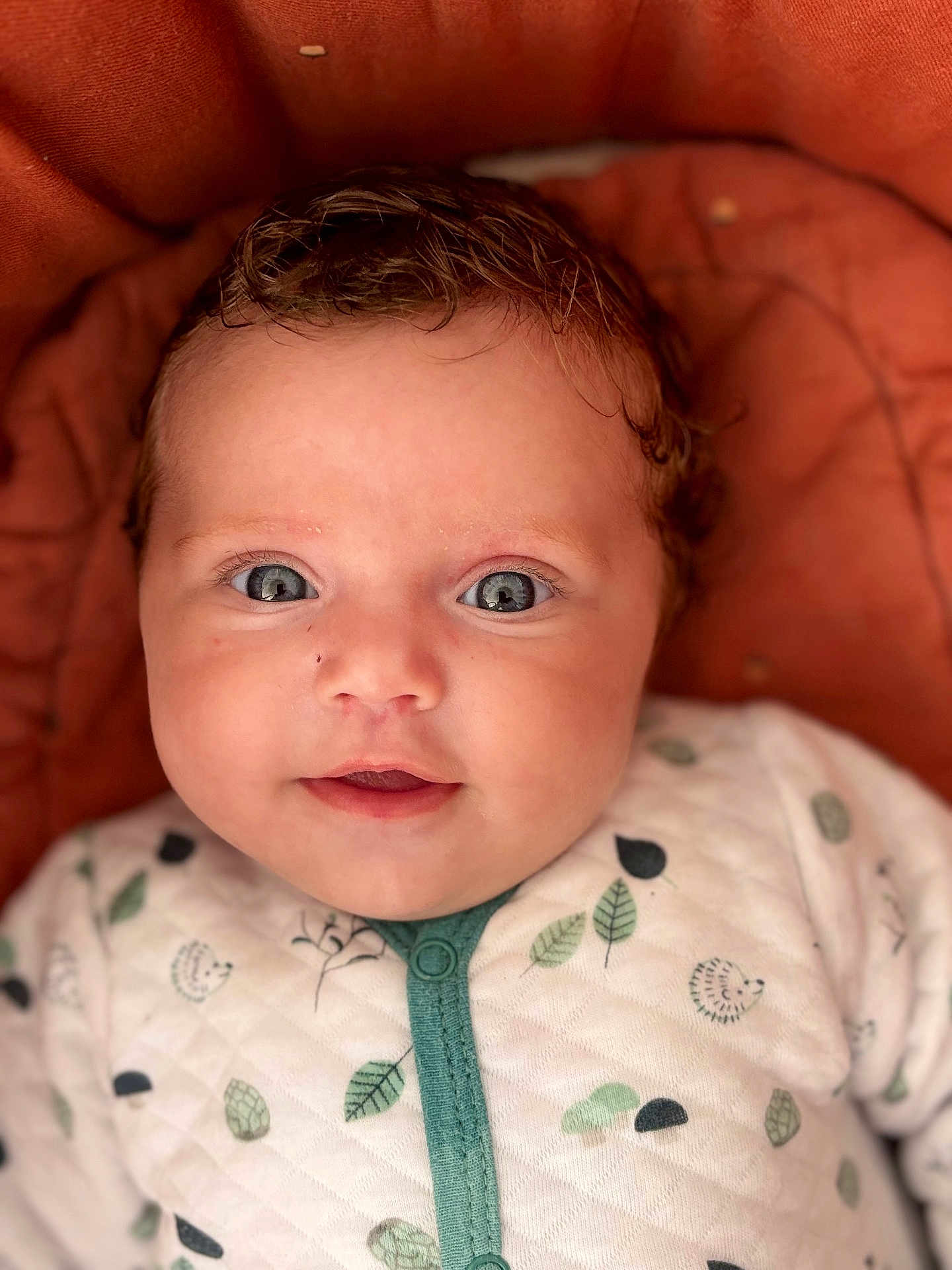 Jaiyanah a rejoint le concours — aidez-le/la à gagner de superbes lots ! baby, infant, face, eyes, curly_hair, onesie, leaf_pattern, hedgehog_pattern, cushion, orange_background, cute, smiling, portrait, closeup, child, soft_lighting, indoors, skin, person, newborn
