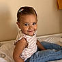 child, toddler, smile, indoor, bed, jeans, striped_shirt, hair_clip, cute, happy, portrait, sitting, skin, face, person, young_child, casual_clothing, neutral_background, soft_lighting, innocence